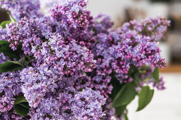 A large purple bouquet of lilacs on the table. In the background is the interior of a modern white Scandinavian-style kitchen. The concept of home comfort and celebration.