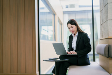 Asian businesswoman sitting in modern office using her laptop and smartphone, enjoying the benefits of a flexible working style.