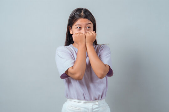 Shy young Asian woman expressing surprise or nervousness with both fists near her mouth, eyes looking sideways. Casual style, isolated on plain background, emotional expression.