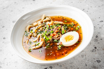 A bowl of ramen with udon noodles, boiled egg, diced vegetables, green onions, and sesame seeds in a rich broth, served in a white bowl on a speckled countertop.