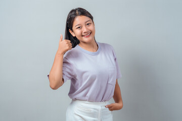Portrait of a cheerful young Asian woman smiling and showing thumbs up while standing against a...