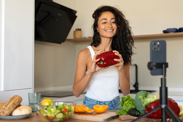 Smiling woman in casual white tank top presenting a red bell pepper to her phone camera while recording a cooking vlog in a bright modern kitchen.