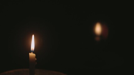 A sharply focused candle flame stands upright in complete darkness. A second distant flame appears as a soft glow behind it. The twisted wax pillar shows dripping edges. The shallow depth of field