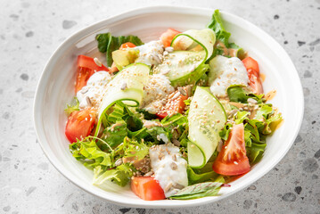 Fresh vegetable salad with tomatoes, cucumbers, lettuce, and seeds, topped with a creamy dressing in a white bowl on a speckled countertop