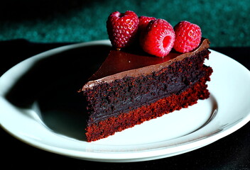 A slice of rich chocolate cake sits on a white plate, adorned with fresh raspberries. The dark cake layers and glossy ganache create a decadent dessert.