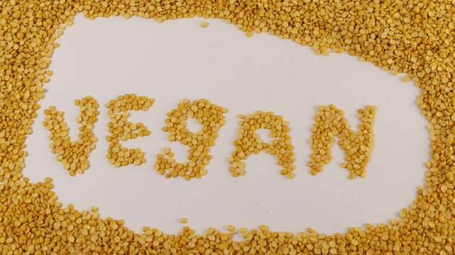 Yellow split peas form the word VEGAN on a seamless white surface. Individual legumes align to create clean letter shapes. Extra peas frame the text in a natural border. Soft overhead lighting softens