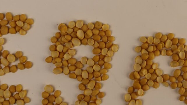 Yellow split peas form the letters VEGAN on a clean white surface. Individual legumes align in precise rows. Extra peas scatter at the top edge. The flatlay composition highlights contrast.