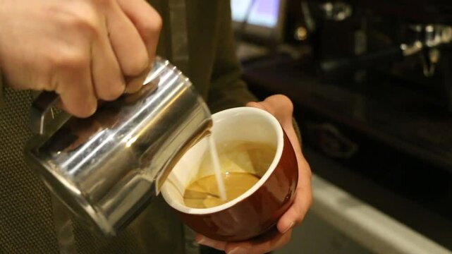 Steamed milk pours into espresso. Ceramic cup fills with creamy layers. Barista crafts latte art heart. Silky microfoam blends with crema. Stainless pitcher tilts precisely. Warm caf&eacute; lighting highlig