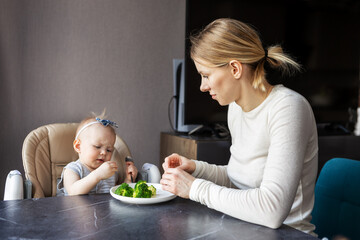 Cute baby girl sitting in high chair and refusing to eat broccoli. Mother Feeding child healthy food. Concept of nutrition, parenting challenges, and child development