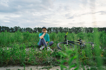 Woman wearing a helmet and sunglasses sitting in a field. She is resting beside her mountain bike....