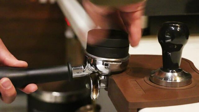 A barista tamps freshly ground coffee in a portafilter. The polished metal tamper presses evenly into dark grounds. A wooden tamp station secures the portafilter. Warm caf&eacute; lighting reflects off chrom