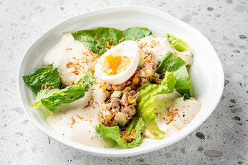 A fresh salad with lettuce, tuna, corn, cucumber, and a halved soft-boiled egg, topped with creamy dressing and crispy fried onions, served in a white bowl on a speckled countertop