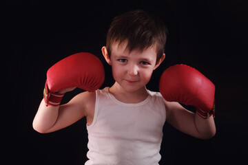 The young fighter, dressed in red gloves and a white shirt, looks serious in front of the dark backdrop. A little boy stands confidently against a black studio background. Kid aged three years