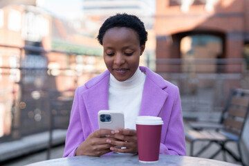 Woman sitting at an outdoor cafe table checking phone messages while enjoying coffee in a city setting on a clear day
