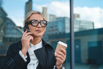 Businesswoman engaged in a phone conversation while holding coffee outdoors in a modern urban...