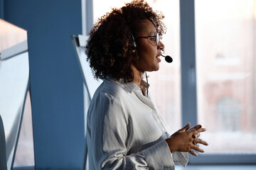 Side view of young African American female expert wearing headset microphone giving presentation at tech conference captured against window in auditorium