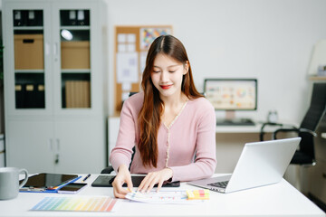 Young graphic designer using pen tablet and laptop at creative desk in modern office. Surrounded by color palettes