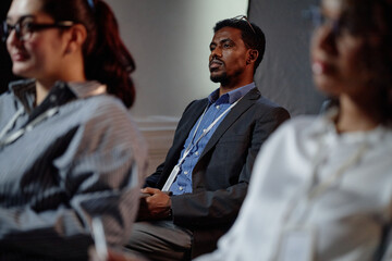 Concentrated Black male manager wearing conference participants badge sitting in audience space with colleagues during executive leadership program meeting
