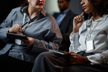 Cropped shot of two female conference participants wearing blank ID badges discussing presenters speech in audience seats during business meeting
