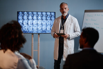 Medium full shot of male head physician holding digital tablet in hands presenting brain MRI scan results displaying on screen at professional conference in auditorium