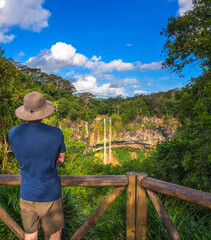 A tourist wearing a hat and casual clothing stands at a wooden viewpoint, looking at the Chamarel Waterfall surrounded by lush greenery in Mauritius.