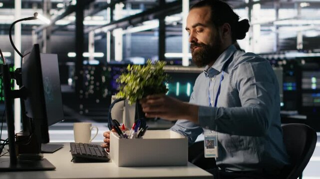 Upset data center worker packing personal items at workstation after being replaced by AI. Programmer removing belongings from server hub workplace desk, fired by artificial intelligence, camera A