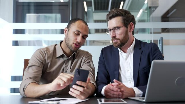 Two male coworkers in office discussing mobile application usage with smartphone. Business man explains to a colleague how to use a new application on a mobile phone, talks and teaches about the app