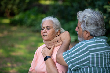 Fototapeta premium Indian senior male supportive collar for the neck to her wife at park. Sick senior woman hurt stiff neck feel fibromyalgia osteoarthritis pinched nerve. Elderly Seniors Health Care living Concept