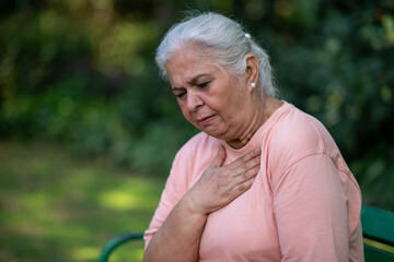 Indian Senior woman holding chest with pained expression sitting on bench outdoors. Possible heart diseases problem, outdoor got troubles with her chest pain or breathing difficulty in park