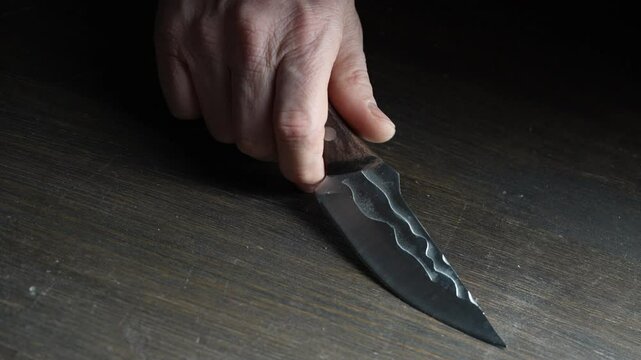 Man's hand takes a forged sharp knife from the wooden table, close up. Iron knife with shiny blade  in male hands