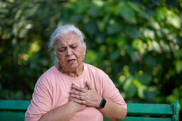 Indian Senior woman holding chest with pained expression sitting on bench outdoors. Possible heart...