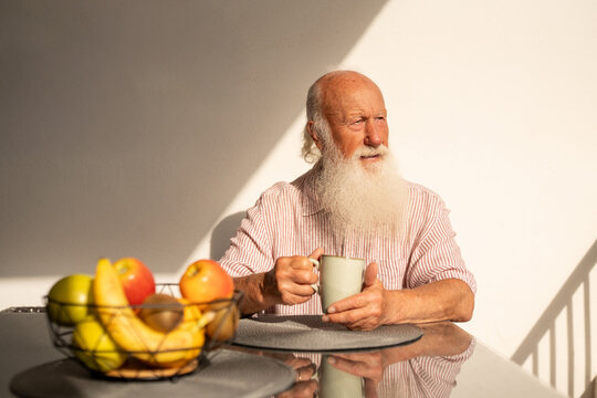 Elderly man with a long white beard sitting at a table with a cup of coffee and bowl of fresh fruits, enjoying sunlight on a peaceful morning