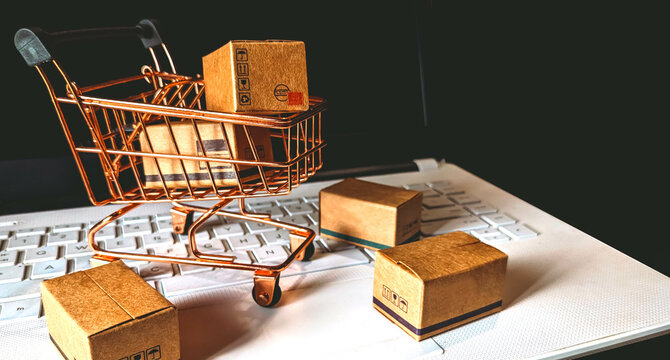 Online Shopping background concept with cardboard and shopping trolley above laptop keyboard on black background - Powered by Adobe