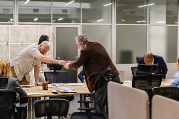 A male employee shakes hands with a male employee standing on the other side of a table with a group of four employees sitting behind it