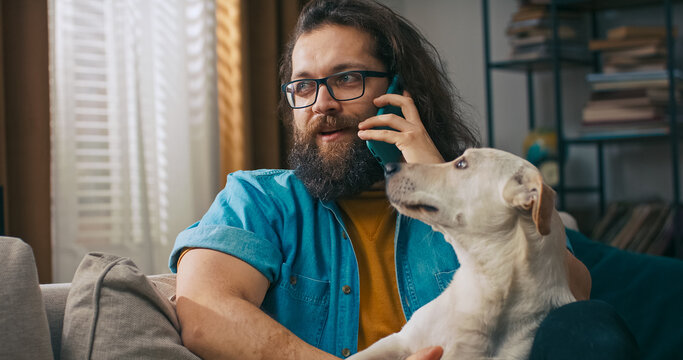 A man holds a phone to his ear and smiles as he speaks, all while his dog rests its head on his lap.
