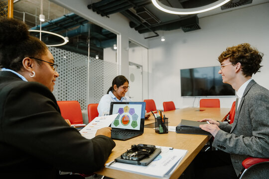 A female worker sits at a desk and holds a paper while looking at a male intern who laughs with a female worker sitting next to her in the office