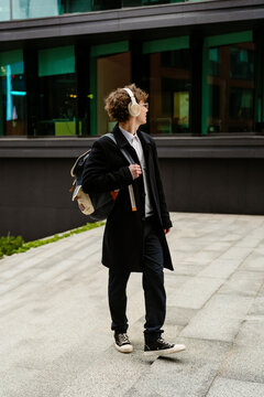 A male intern in headphones walks and looks away while holding a backpack on his shoulder, on the street