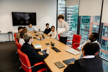 A group of nine employees sit at a table and listen to a male intern standing next to them talking...