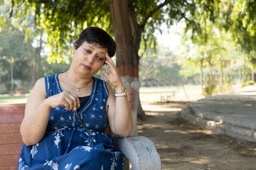 Indian worried woman sitting alone on bench look down feel sorrow touching head brain at park....