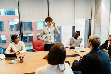 Male intern standing and holding clipboards while talking to a group of five employees sitting at a table in an office