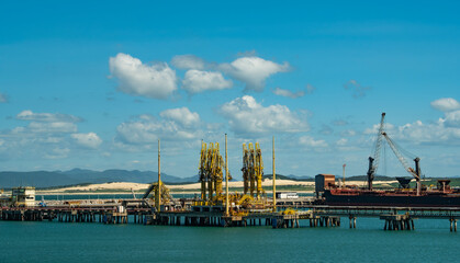 Obraz premium Busy port scene. Large cargo ship docked at industrial pier with cranes under a bright blue sky with scattered clouds