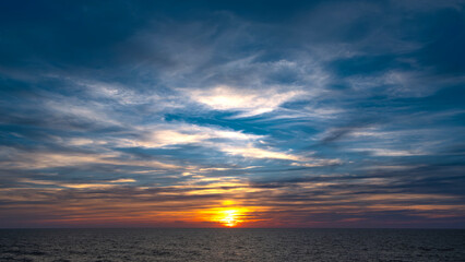 Obraz premium Dramatic sunset over the ocean. Vibrant colors paint the sky with clouds reflecting golden light on the water. Peaceful, serene scene
