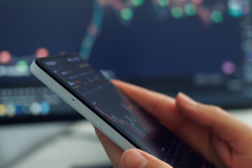 User interacts with stock market data displayed on a smartphone, with market analytics visible on a computer screen in the background