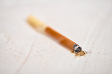 Close-up of cigarette resting against white fabric, leaving burn mark. Soft lighting highlights damage, contrasting with brown and white tones. Shallow depth of field focuses on burn area