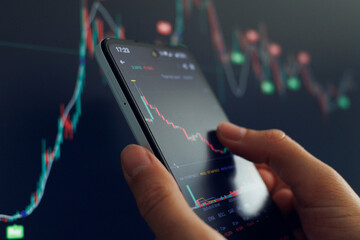 A person examines stock market trends on a smartphone, with colorful charts and data visible,...
