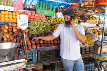 Indian shopkeeper man selling organic veggies and fruits on a street market while talking on phone
