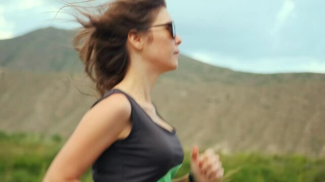 Side view. A fit woman in sunglasses runs along a deserted road, with mountains and green hills behind. Her fast pace and steady breath reflect endurance and commitment to regular training.