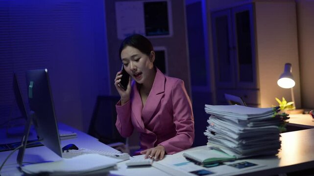 A woman is talking on her cell phone while sitting at a desk with stacks of papers. She is wearing a pink jacket and she is in a happy mood