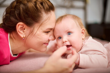 Young woman with brown hair and smiling baby girl on pink bed. Light-hearted interaction, creating joyful atmosphere. Warm lighting enhances bond