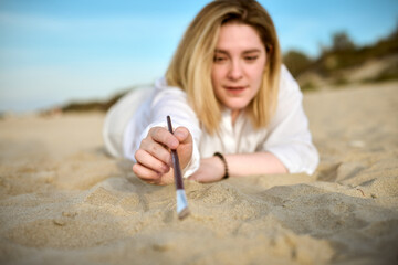 Young woman uses paintbrush on sandy beach. Serene coastal environment and soft sunlight evoke creativity and relaxation. Her delicate touch on sand suggests moment of artistic inspiration